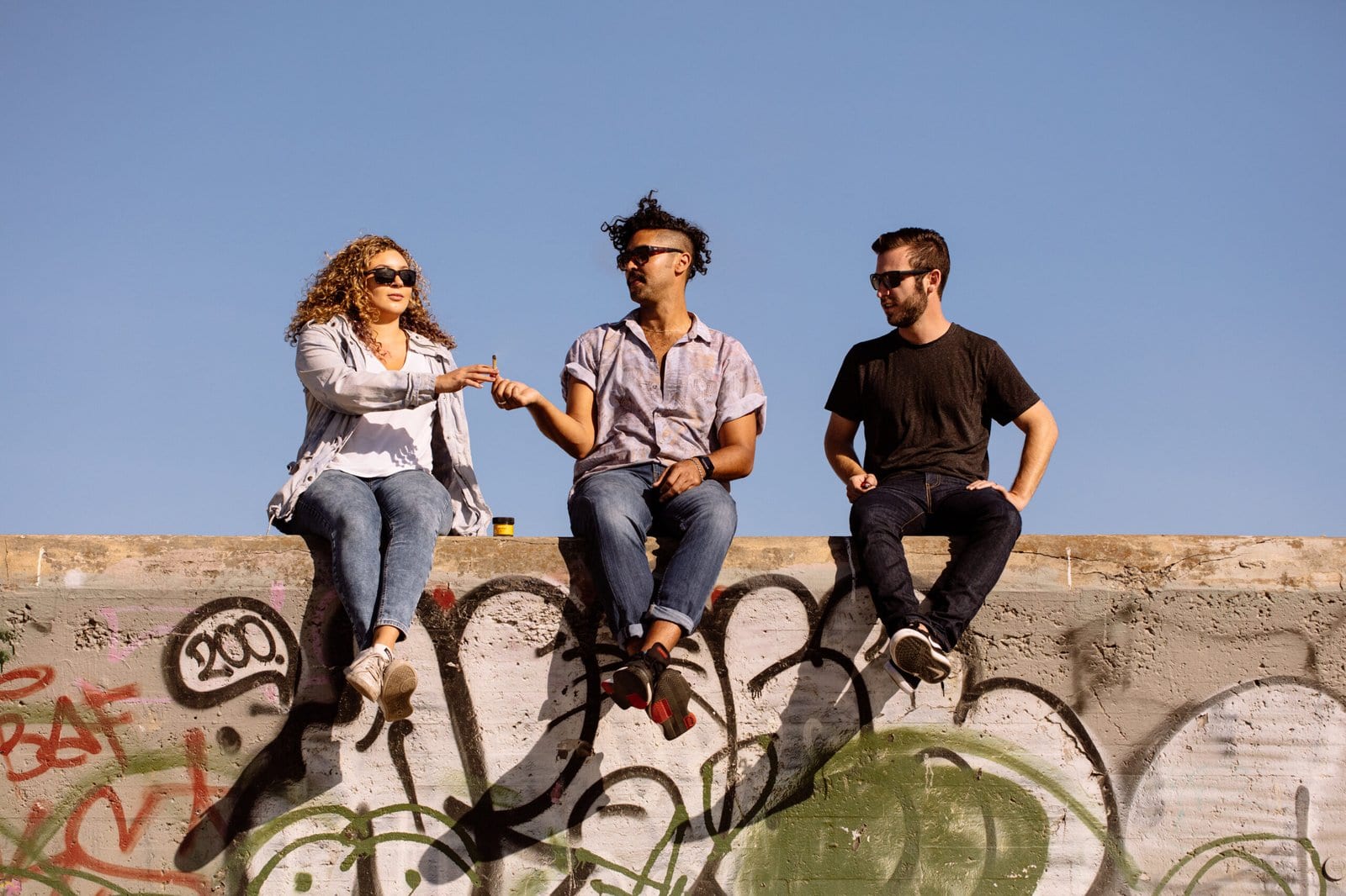 Three friends passing a joint sitting on top of a concrete ledge that's covered in graffiti