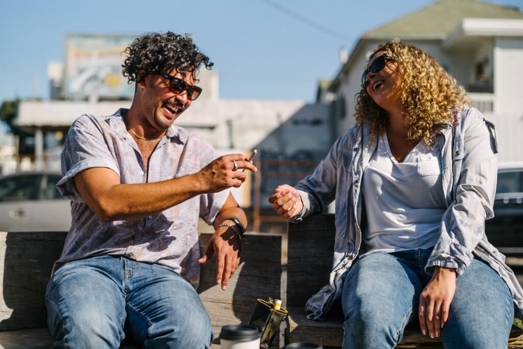 A man and woman laughing and passing a joint while sitting on a bench near the roadside.
