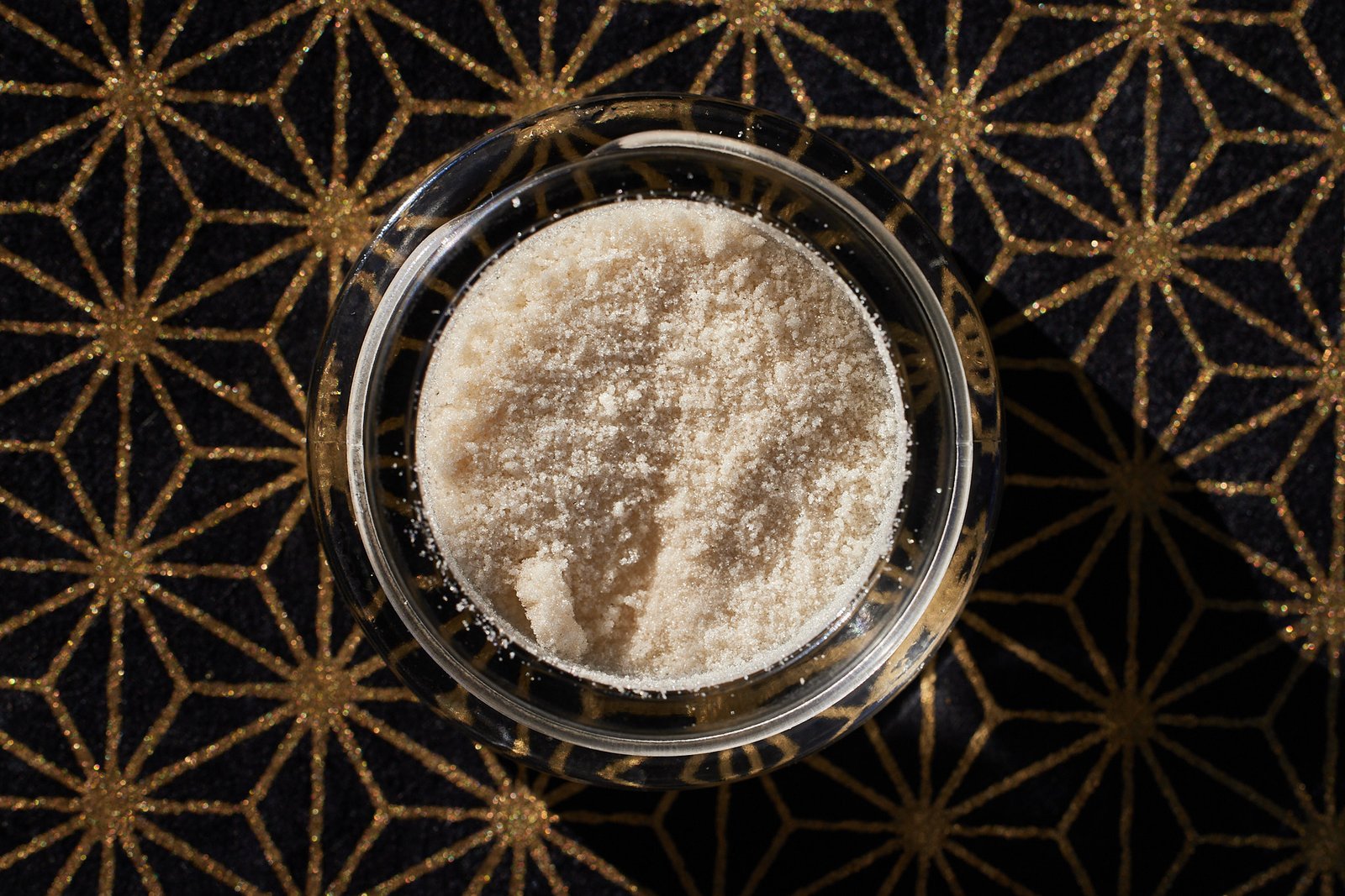 Jar of full melt hash sitting on gold and black geometric pattern background with hard light casting a shadow from the side of the jar